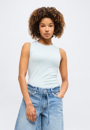 Νεαρή γυναίκα με σγουρά μαλλιά όρθια, φοράει αμάνικη ανοιχτό μπλε υφασμάτινη μπλούζα και φαρδιά ανοιχτόχρωμα τζιν με δεμένη ζώνη.