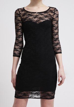 Femme portant une robe en dentelle noire avec des manches longues transparentes et la partie supérieure de la poitrine, silhouette ajustée et ourlet au-dessus du genou.