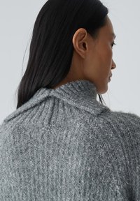 Grau gestrickter Pullover mit hohem Kragen und geripptem Muster, das lose Fäden enthält, die durch den Stoff gewoben sind.