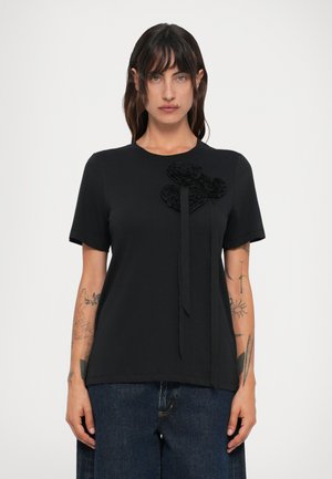 Woman with shoulder-length dark hair wearing a black short-sleeve top with 3D floral detail and blue jeans, standing against a white background.