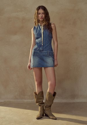 Mladá žena s dlhými vlnitými vlasmi má na sebe bezrukávové rifľové šaty a opálené voľné kovbojské topánky, stojaca pred jednoduchým béžovým pozadím.