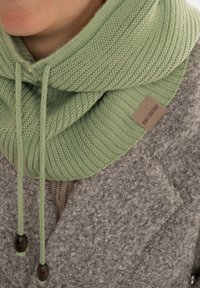 Gebreide groene kraag met een ribbelstructuur en trekkoorden, met houten toggles. Draagt over een lichtgrijze, fuzzy jas. Kleine label aanwezig.