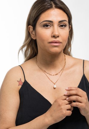Goldene Schichtenketten mit einem großen Perlenanhänger, getragen über einem schwarzen ärmellosen Oberteil. Das Modell hat ein Sterntattoo auf ihrer Schulter.