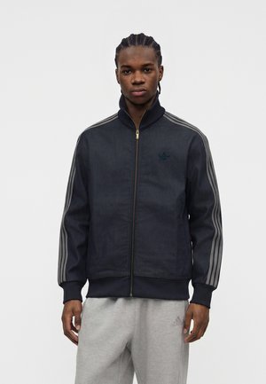 Ung mand med fletninger iført en mørk lynlåstrøje med grå striber på ærmerne og lysegrå Adidas træningsbukser.