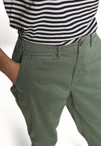 Zelené bavlněné chinos kalhoty se rovný fit. Disponují přední kapsou, poutky na opasek a decentním šitím. Jsou kombinovány s pruhovaným černobílým topem.