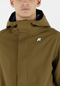 Giacca impermeabile verde oliva con colletto alto, trama sottile e un piccolo logo metallico sul lato sinistro del petto.