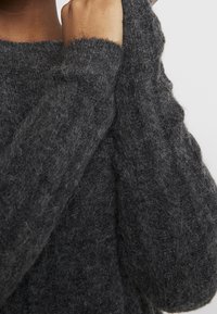 Maglione grigio scuro lavorato a maglia con una finitura texturizzata e pelosa. Presenta dettagli a coste sulle maniche e un collo rotondo. Primo piano sul tessuto e sui punti.