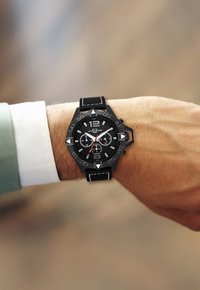 Reloj cronógrafo negro con esfera texturizada en negro, acentos blancos, marcadores triangulares y correa de cuero. Lleva puesto en una muñeca con una camisa verde claro.