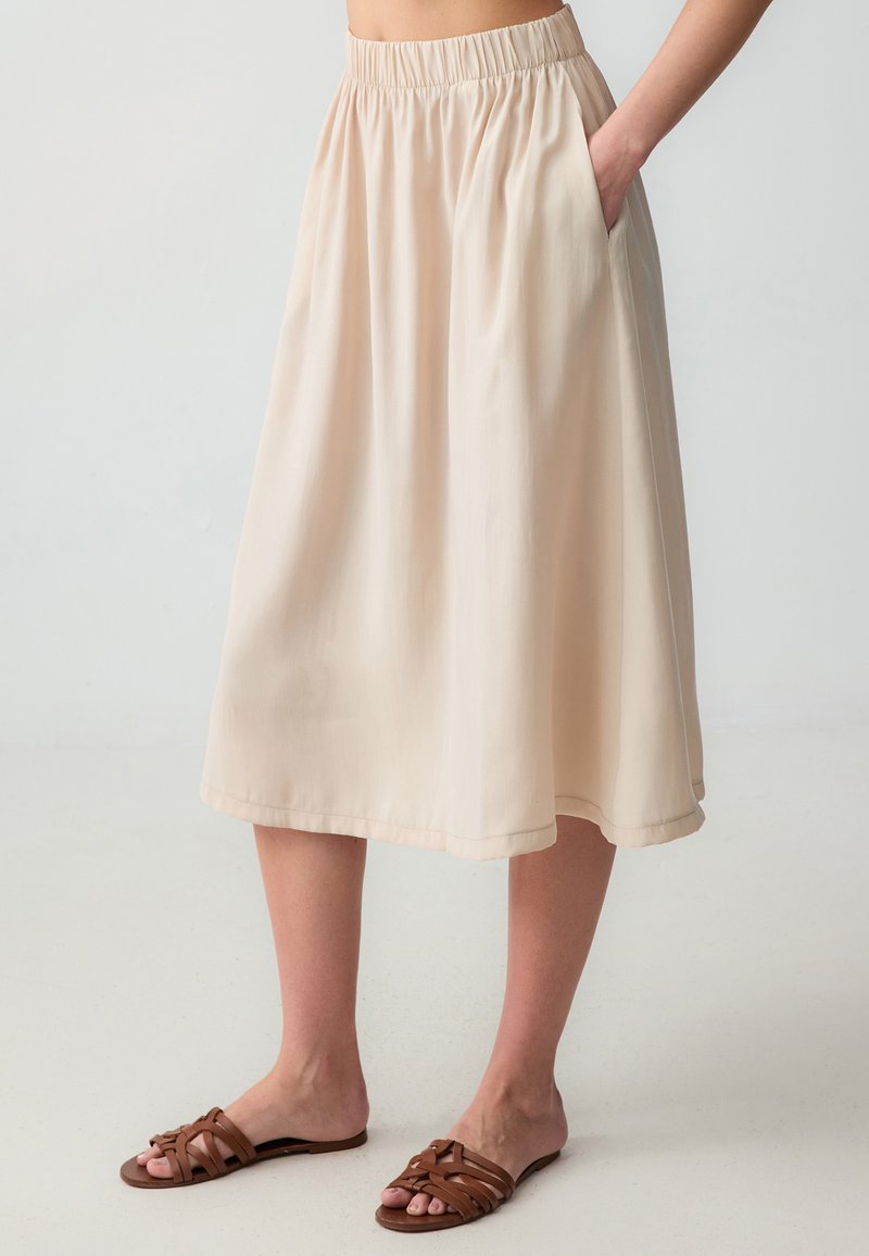 Jupe midi beige clair avec une taille élastique et des poches latérales inclinées. Le tissu lisse drape délicatement, l'ourlet tombant juste en dessous du genou.