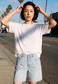Jeune femme ajuste ses cheveux, portant un T-shirt blanc et un short en jean clair, debout dans une rue urbaine ensoleillée avec des palmiers.
