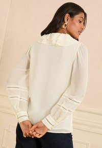 Hvid bluse med flæset krave og gennemsigtige lange ærmer. Har dekorative udsparinger på ærmerne og knaplukninger ved manchetterne. Enkel, elegant design.
