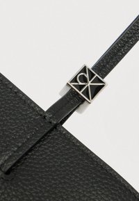 Μαύρο αξεσουάρ από υφή δέρματος με ορθογώνιο ασημί και μαύρο λογότυπο, που διαθέτει κομψό σχεδιασμό λουριού για ασφαλή κλείσιμο.