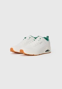 Par med hvite sneakers med grønn innvendig fôr og oransje gummisåler, med synlig luftdemping og snøring.