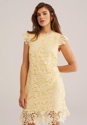 Frau mit braunen Haaren, die ein blassgelbes, ärmelloses Spitzenkleid mit floralen Mustern und geschwungenen Rändern trägt, steht vor einem schlichten Hintergrund.