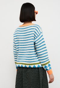 Femme aux cheveux courts et foncés portant des lunettes orange et un pull rayé bleu et blanc avec des motifs géométriques, tournée dos à la caméra.