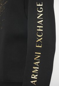 Μαύρο φούτερ με χρυσό κείμενο "ARMANI EXCHANGE" κατά μήκος του μανικιού, με λεία υφή και μινιμαλιστικό σχέδιο.