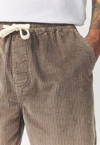 Braune Cordhosen mit vertikal geripptem Muster, ausgestattet mit einem elastischen Bund, einer Kordelzugschliee und Seitentaschen.