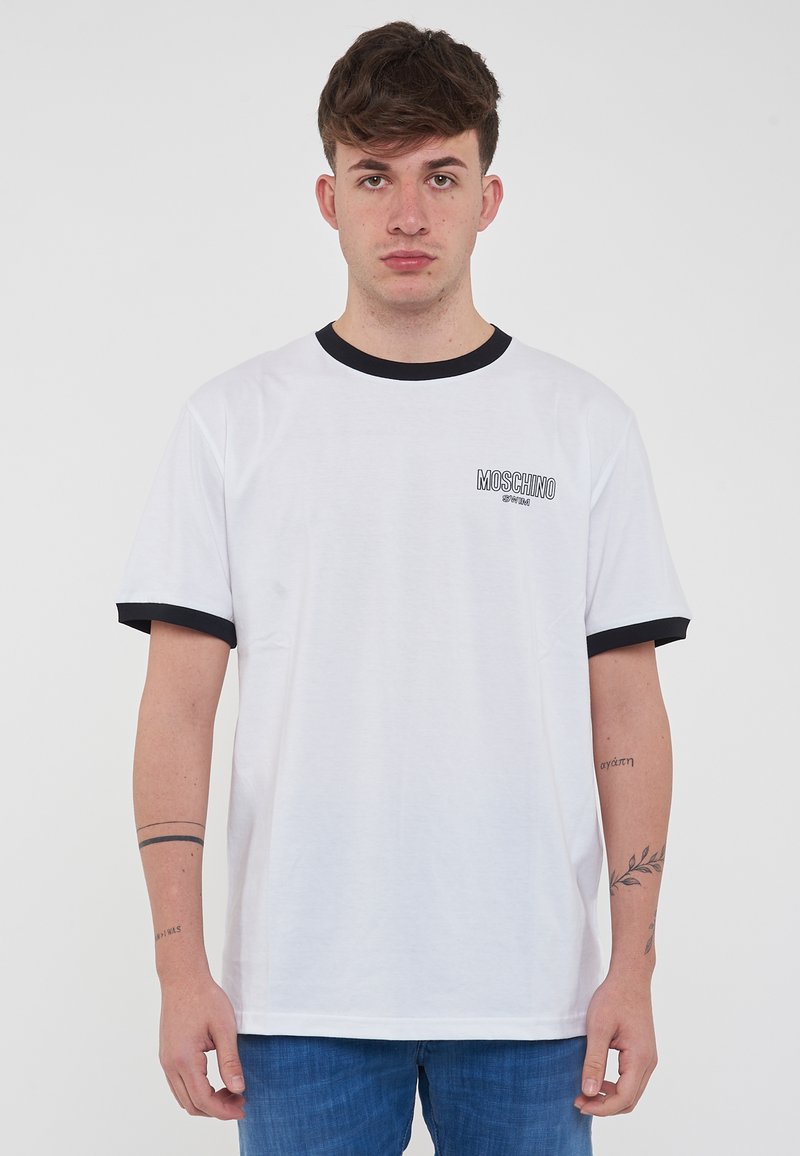 Maglietta bianca in cotone con collo e polsini neri a costine, dotata di un piccolo logo nero sul lato sinistro del petto. Vestibilità classica con maniche corte.