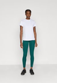 Weißes kurzärmliges T-Shirt kombiniert mit türkisfarbenen Leggings mit reflektierenden Akzenten und schwarzen Sportschuhen. Das Modell steht vor einem einfarbigen Hintergrund.