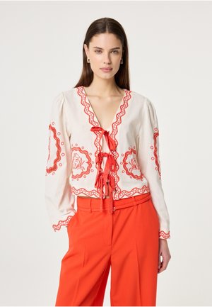 Frau trägt eine weiße Bluse mit roter Blumenstickerei und Bändern, kombiniert mit hoch taillierter roter Hose, steht vor einem schlichten Hintergrund.