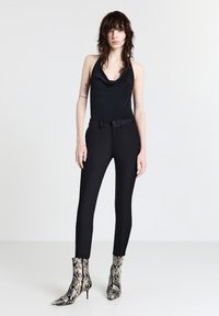 Femme aux cheveux mi-longs portant un haut sans manches noir, un pantalon noir ajusté et des bottines à motif serpent, se tenant devant un fond blanc.