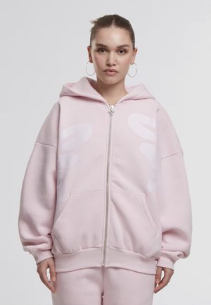 Frau mit braunem zurückgebundenem Haar, die große Creolen und einen hellrosa oversized Zip-Hoodie sowie passende Hose trägt, blickt nach vorne.