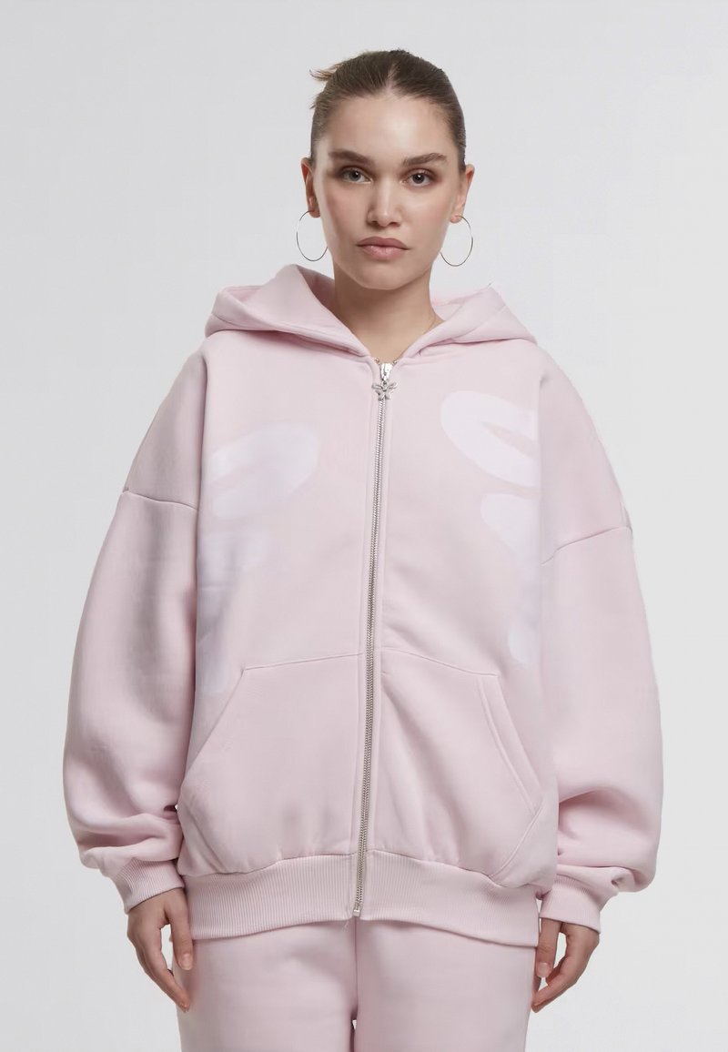 Femme aux cheveux bruns attachés, portant de grandes boucles d'oreilles créoles et un sweat à capuche zippé rose pâle oversize avec un pantalon assorti, de face.