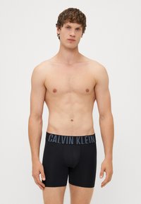 Boxeri negri cu talie lată, având inscripția "CALVIN KLEIN" în albastru închis. Textură netedă, potrivire ajustată și design liniar.