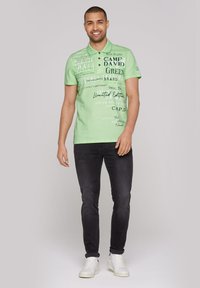 Zelená polo tričko s textúrovanou tkaninou, s rôznorodou typografiou a logami. V kombinácii s tmavými slim-fit džínsy a bielymi teniskami.