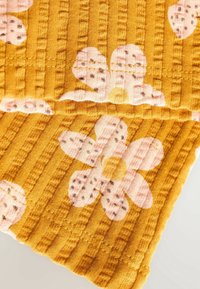 Tissu jaune texturé avec des côtes surélevées, présentant de grandes fleurs roses avec des taches foncées. Détail des coutures visible le long du bord.