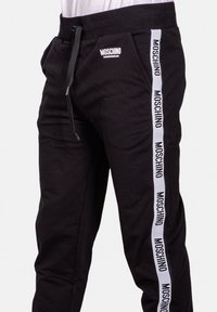 Svarta sweatpants med dragsko, sidofickor och ett vertikalt vit logostrimma där märkesnamnet "Moschino" upprepas.