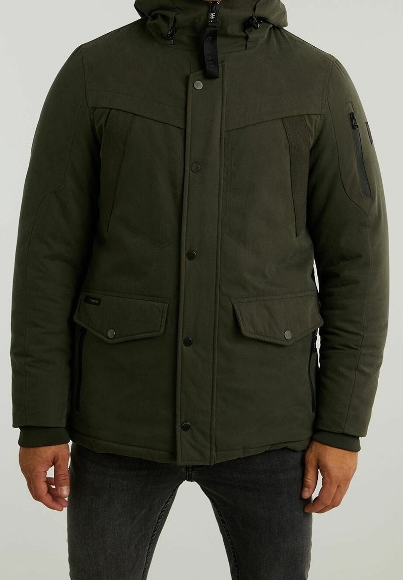 Hombre con chaqueta de invierno con capucha de color verde oliva, con bolsillos delanteros con solapa y cierres de botones, combinado con jeans gris oscuro.