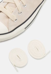 Beige suede sneaker with white rubber toe cap and black sole, featuring beige laces threaded through silver eyelets. Two white laces beside.