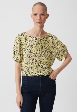 Blouse fleurie jaune, avec des manches courtes et un large décolleté. Le tissu a une texture avec des motifs noirs et violets éparpillés.