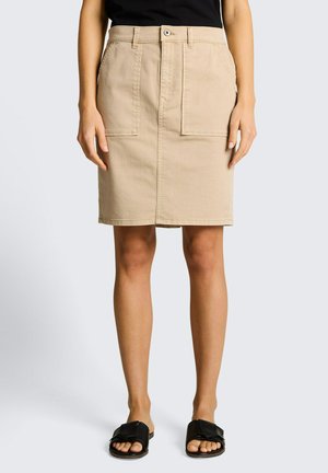 Jupe beige mi-longue avec grandes poches avant, fermeture par bouton et zip, portée avec un haut noir et des sandales noires à bout ouvert.