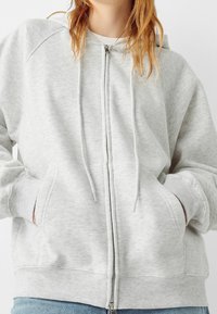 Felpa con zip grigia chiara, dal texture morbido, con polsini a costine e tasche anteriori. Presenta un cappuccio con coulisse e una chiusura con zip.