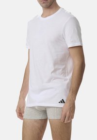 White crew neck t-shirt made of soft cotton, features a small black Adidas logo on the lower left side. Worn with gray fitted shorts.