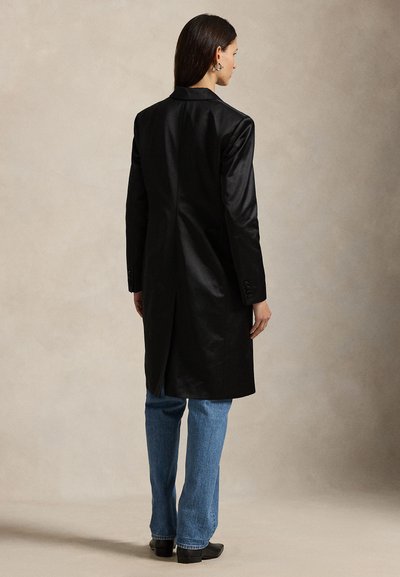 Casaco longo de cetim preto com gola de lapela, abertura atrás e punhos com botões, usado sobre jeans azuis e botas de salto. Fundo claro simples.