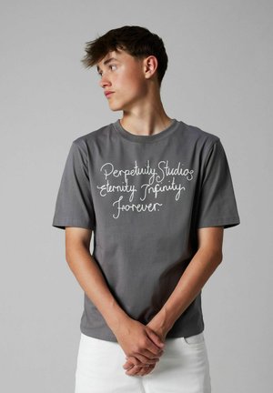 Giovane uomo con t-shirt grigia con testo bianco "Perpetuity Studios eternity Infinity forever" che guarda a sinistra, con le mani intrecciate davanti, su uno sfondo neutro.