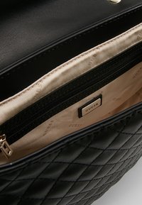 Černá prošívaná kabelka s hladkým, lesklým povrchem a přihrádkou na zip. Vnitřek je vyložen béžovou tkaninou s logovou visačkou.