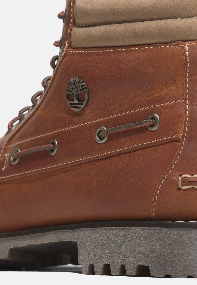 Brązowy skórzany but z beżową cholewką, metalowymi oczkami, sznurówkami oraz teksturowaną gumową podeszwą z detalem w postaci logo drzewa.