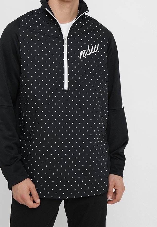 Homme portant un pull noir à manches longues avec des pois blancs, une fermeture éclair blanche et le texte blanc "nsw" sur la poitrine, debout devant un fond uni.