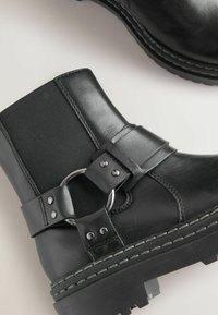 Sorte ankle boots lavet af læder med et stofpanel, der har et cirkulært metalaccent, stropdetalje og tekstureret sål.