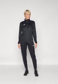 Svart idrettsdrakt med lange ermer og høy krage. Har hvite striper på ermer og bukser, Adidas-logo på brystet og hoften.