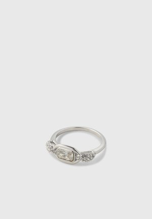 Bague en argent avec une pierre précieuse rectangulaire claire au centre, flanquée de petites pierres rondes claires de chaque côté, sur un fond uni.
