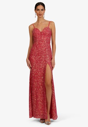 Frau trägt funkelndes rotes bodenlanges Kleid mit dünnen Trägern und hohem Schlitz, kombiniert mit hängenden Ohrringen und spitzen High Heels.