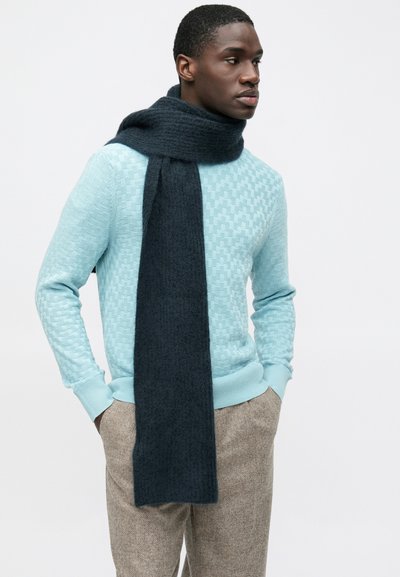 Pull tricoté bleu clair avec un motif texturé, associé à une écharpe gris foncé drapée autour du cou. Pantalon aux tons neutres.