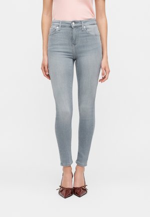 Kvinde iført lysegrå skinny jeans, lyserød top og burgundy spidse hæle, der står mod en ensfarvet hvid baggrund.