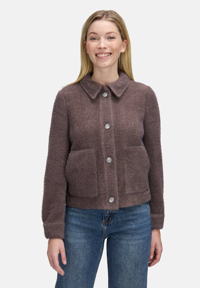 Brun, tekstureret, kort jakke med krave, fem knapper og to frontlommer. Båret over en sort top og blå jeans.