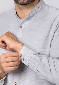Hellgrau kariertes Hemd mit Kragen, ausgestattet mit braunen Knöpfen und einer Lasche am Bündchen. Der Stoff wirkt strukturiert und atmungsaktiv.